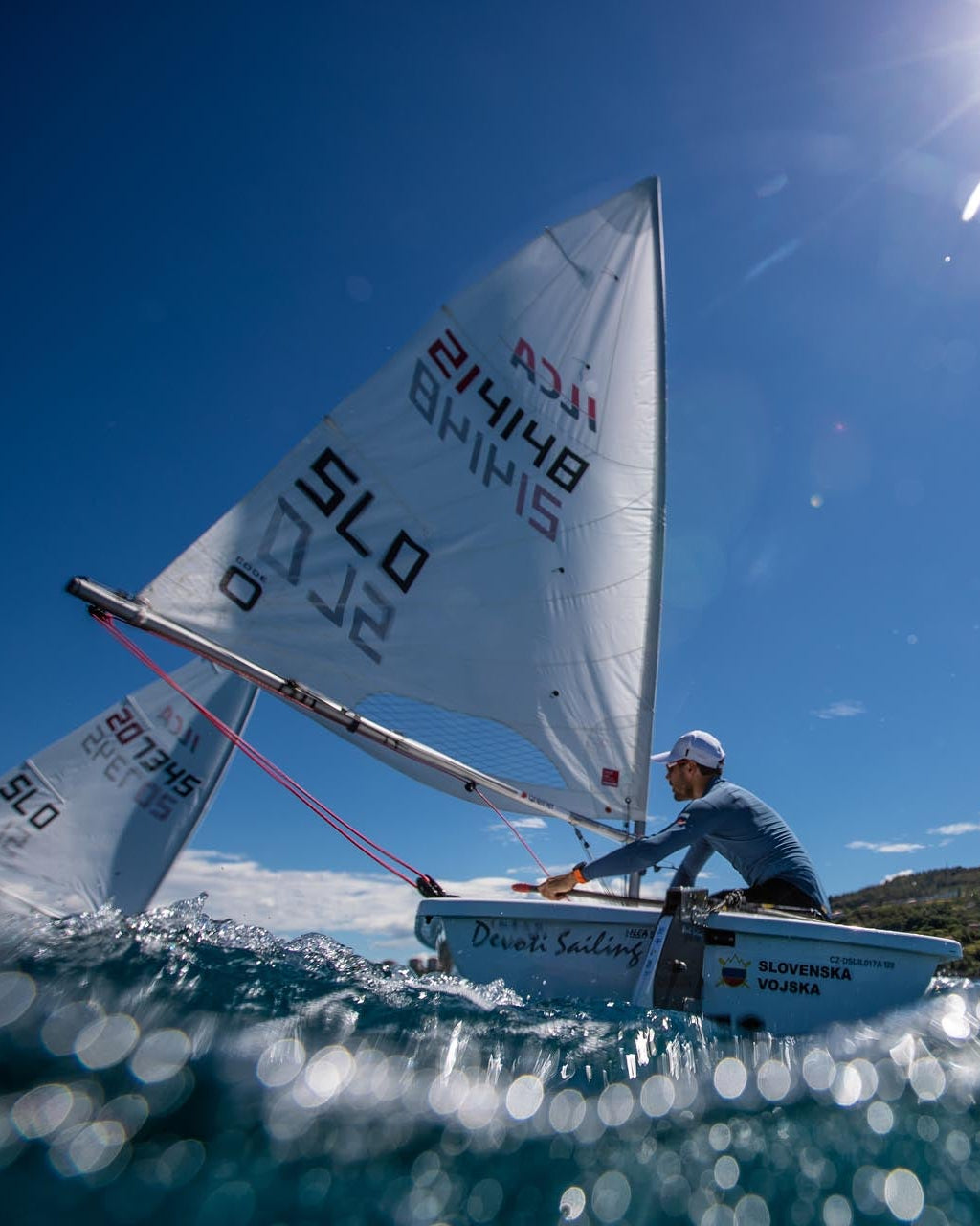 A person sailing a small white dinghy on blue water under a bright sun and clear blue sky, with hills visible in the distance. The sail has visible text including 'SLO' and 'Briant'.
